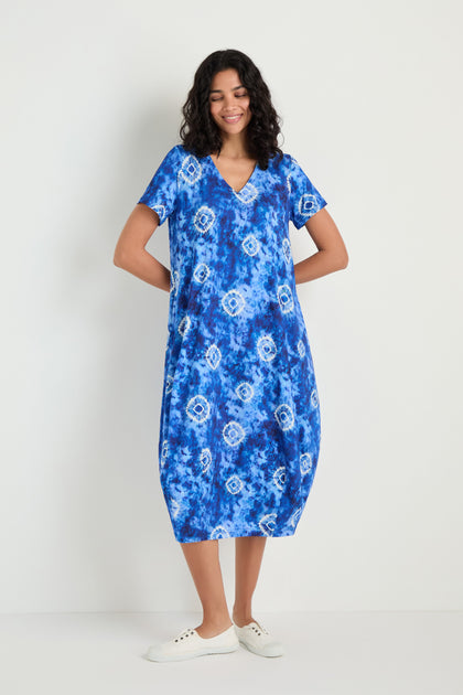 A woman poses against a white background wearing the Tie Dye Circles Jersey Bubble Dress in blue viscose stretch jersey, featuring short sleeves, a knee-length hem, and styled with white sneakers.