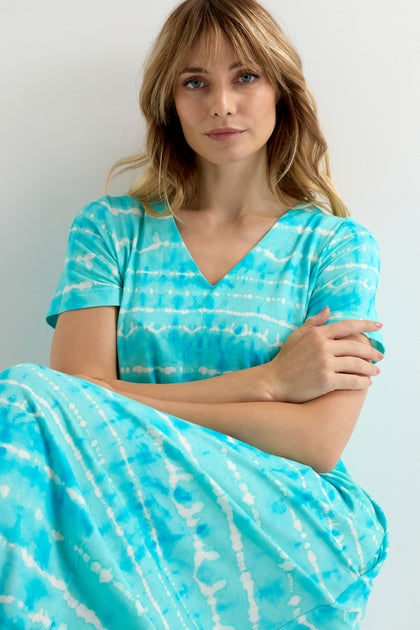 Woman with wavy blonde hair wears the Turquoise Tie Dye Jersey Bubble Dress, sitting with folded arms and looking at the camera against a plain light background.