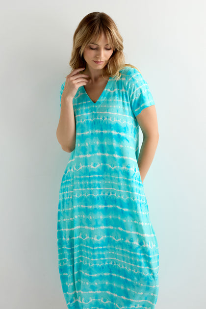 A woman stands against a light background wearing the Turquoise Tie Dye Jersey Bubble Dress, featuring a chic bubble silhouette and V-neck in sea glass-inspired shades of blue and white.