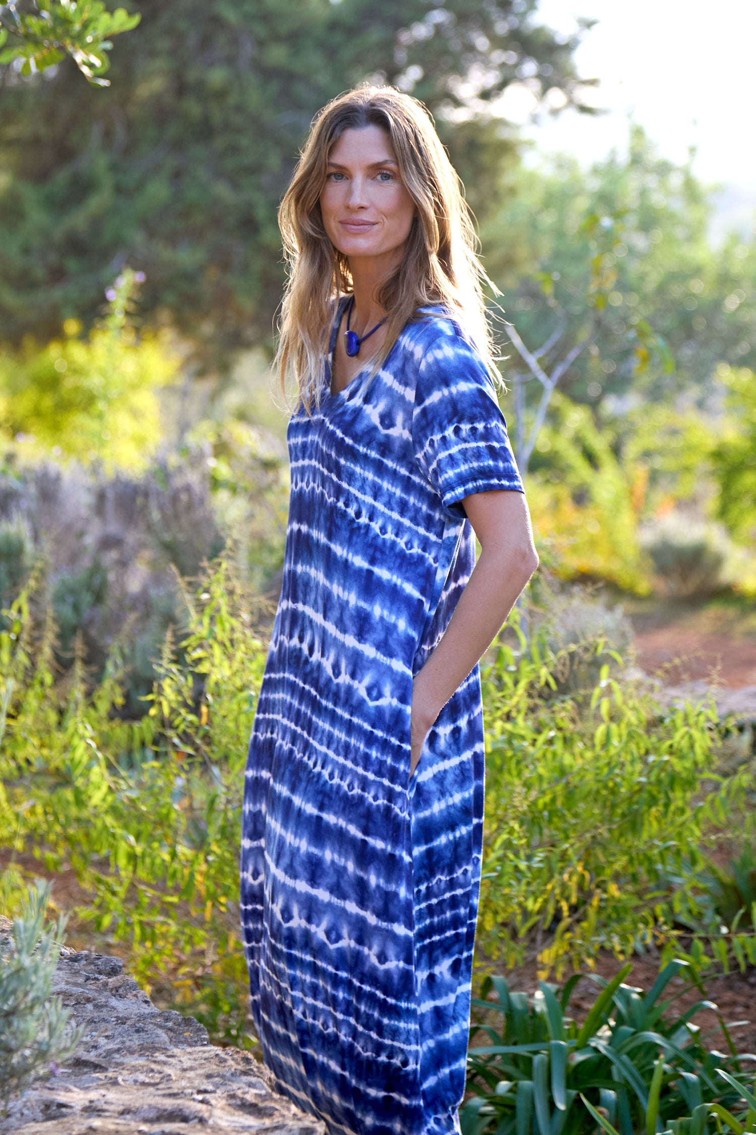 Amidst lush greenery, the woman confidently gazes at the camera, wearing a blue Tie Dye Jersey Bubble Dress.