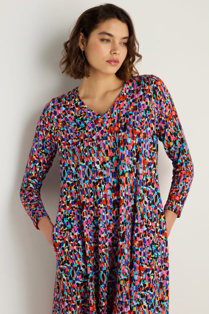 Woman models the Painted Spots Jersey Flared Dress—midi length with multicolored abstract spots and pockets—standing against a plain, light background.
