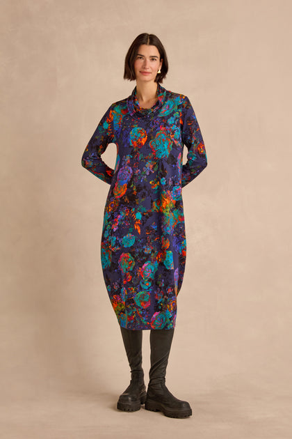 A woman stands against a neutral background, wearing the Midnight Floral Jersey Bubble Dress in a midi length with a colorful abstract pattern, paired with black boots.