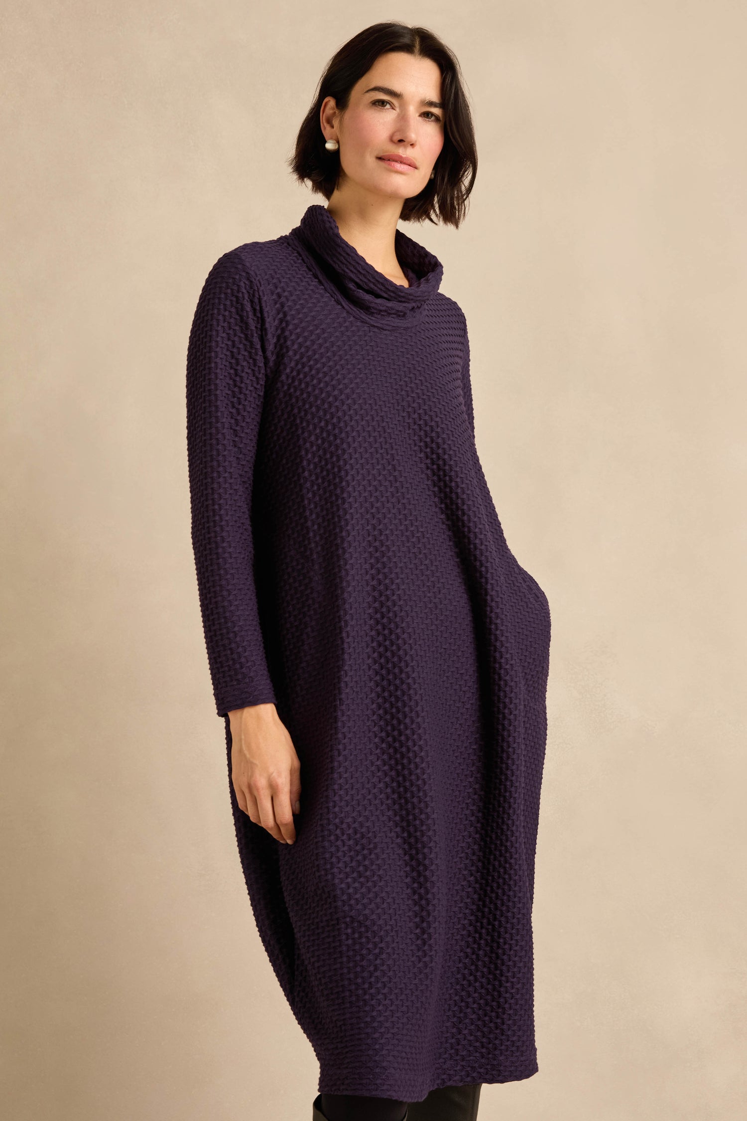 A woman stands against a beige background with one hand in her pocket, wearing the Damson Waffle Textured Jersey Bubble Dress, a long-sleeve purple cowl neck style.