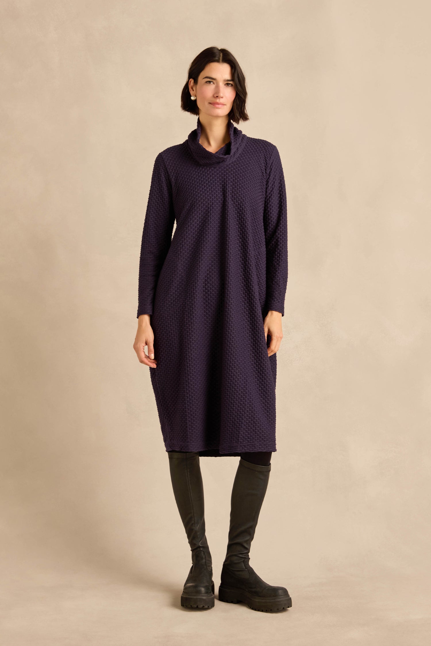 A person stands against a beige background wearing the Damson Waffle Textured Jersey Bubble Dress—a long-sleeve, knee-length bubble dress in purple waffle-textured jersey—paired with black boots.