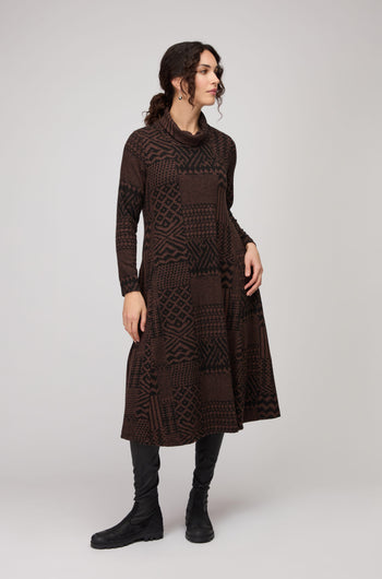 Woman in a studio wears the Textured Ikat Patchwork Jersey Dress in brown and black with long sleeves, styled with black knee-high boots.