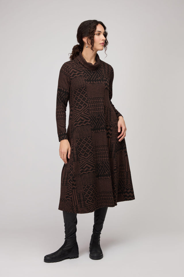 Woman in a studio wears the Textured Ikat Patchwork Jersey Dress in brown and black with long sleeves, styled with black knee-high boots.
