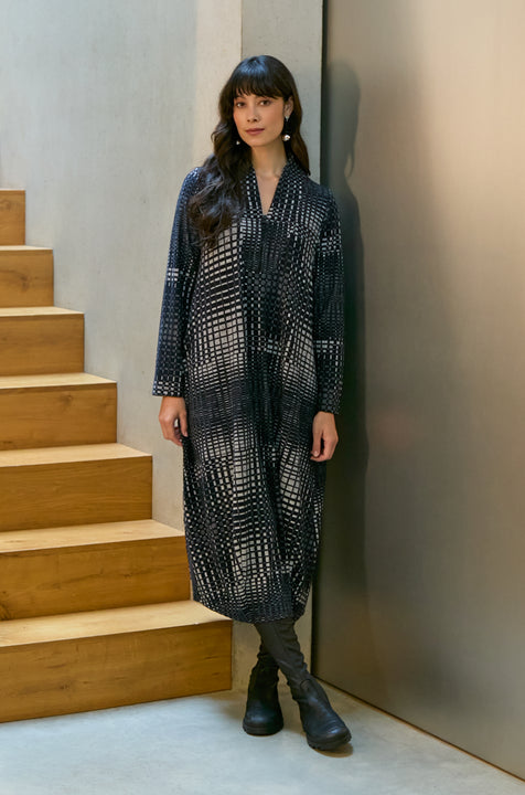 A woman stands indoors by wooden stairs, wearing the Illusion Check Jersey Bubble Dress and black boots, with her hair down and hands by her sides.