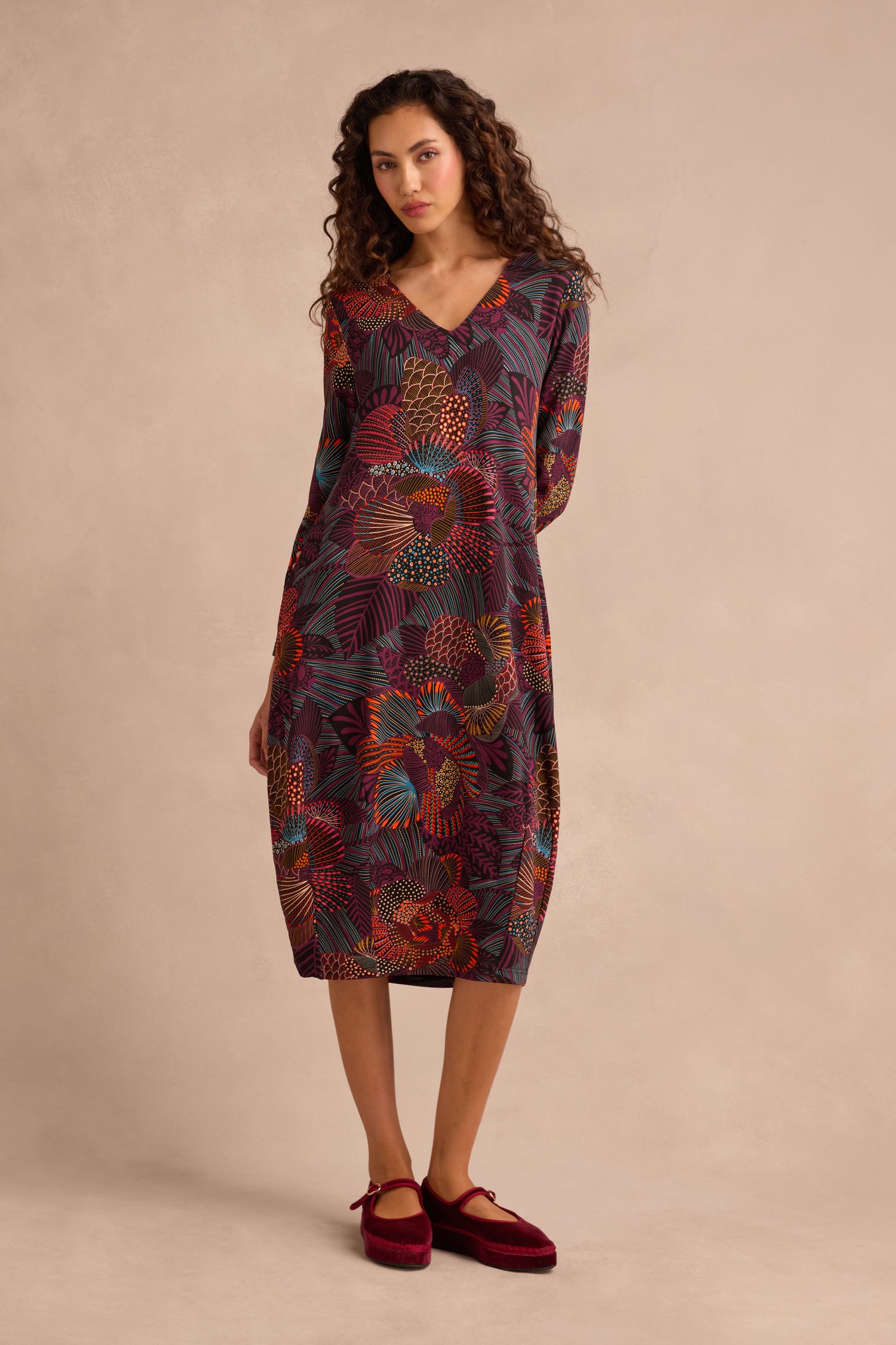 A woman stands against a neutral background wearing the Abstract Linear Floral Jersey Bubble Dress—a long-sleeved, knee-length dress featuring a colorful abstract leaf pattern. Her arms rest naturally at her sides, and she completes the look with red shoes.