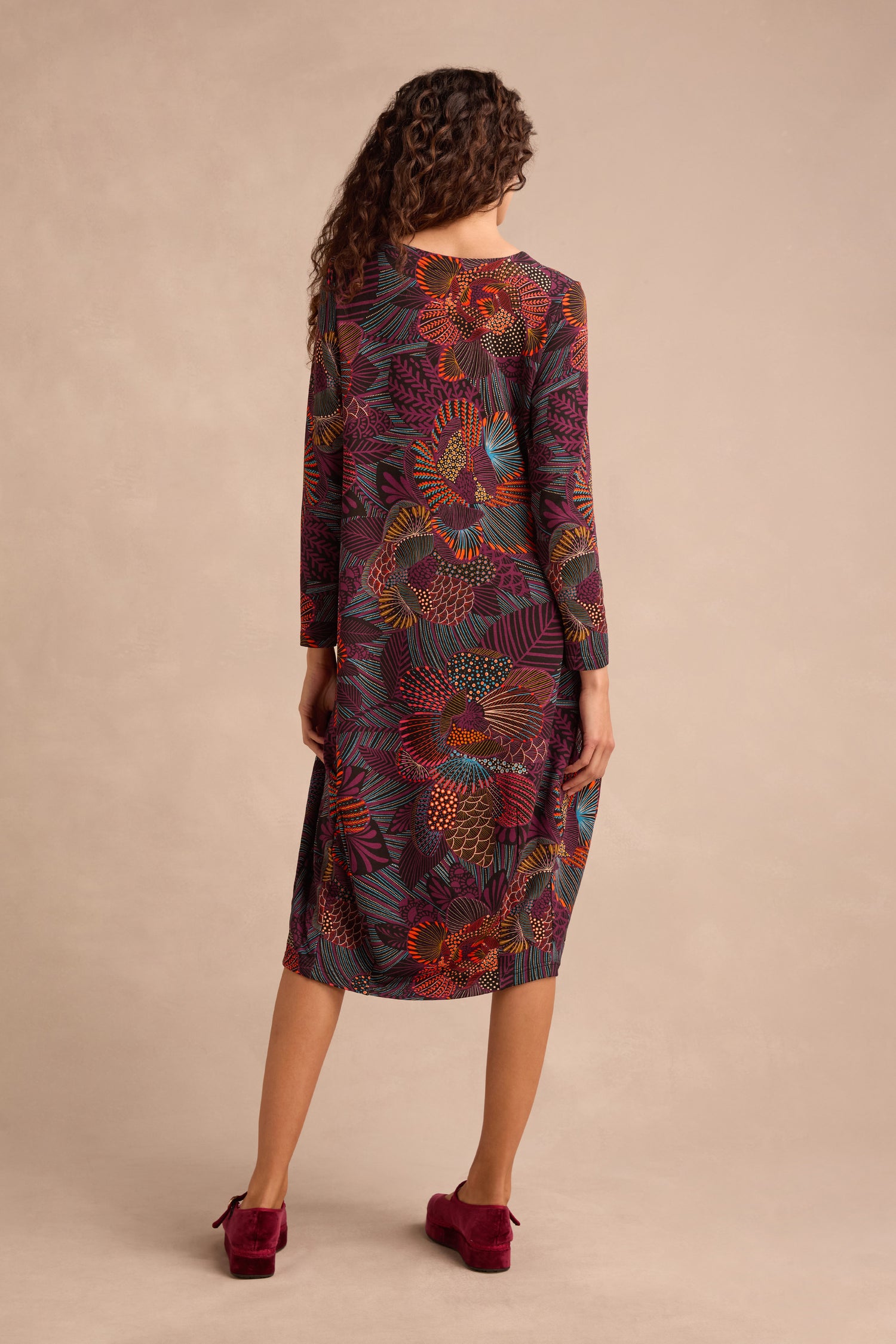 A woman stands facing away against a plain beige background, wearing the Abstract Linear Floral Jersey Bubble Dress—a long-sleeved, colorful floral midi dress—paired with red shoes.