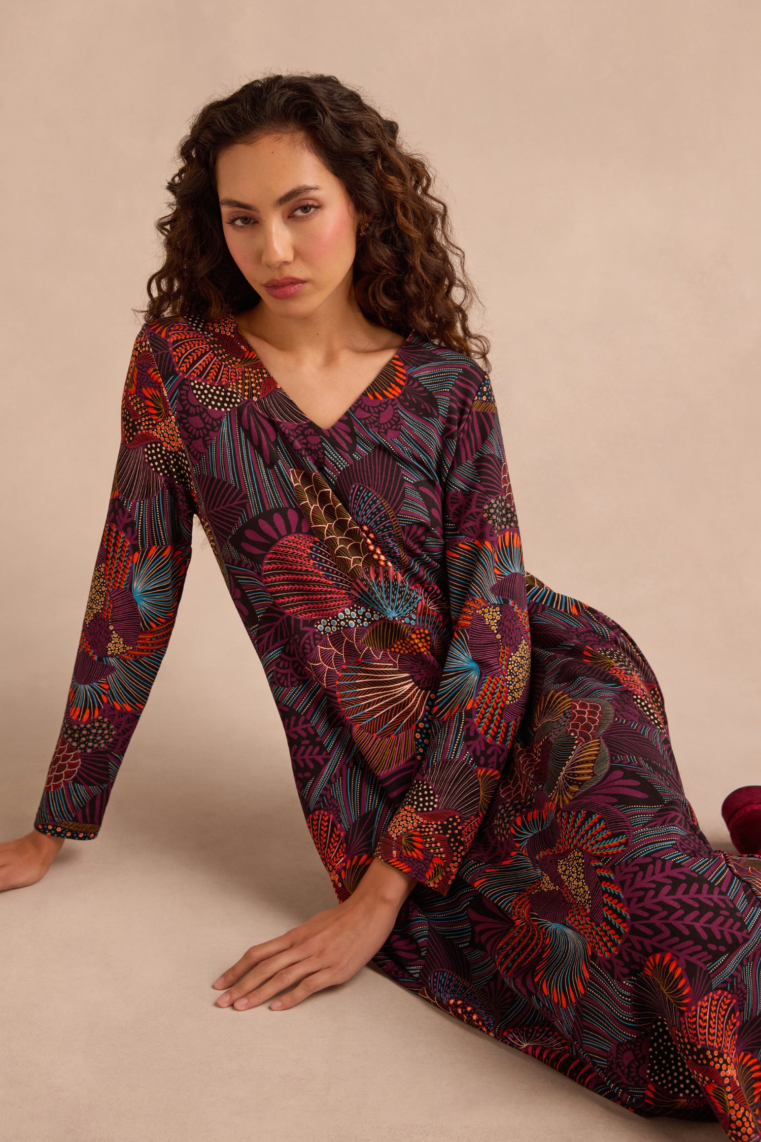 A woman with long, curly hair wears the Abstract Linear Floral Jersey Bubble Dress—a long-sleeve, multicolored floral piece—and sits on the floor against a plain beige background.