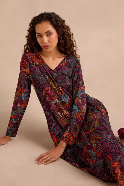 A woman with long, curly hair wears the Abstract Linear Floral Jersey Bubble Dress—a long-sleeve, multicolored floral piece—and sits on the floor against a plain beige background.