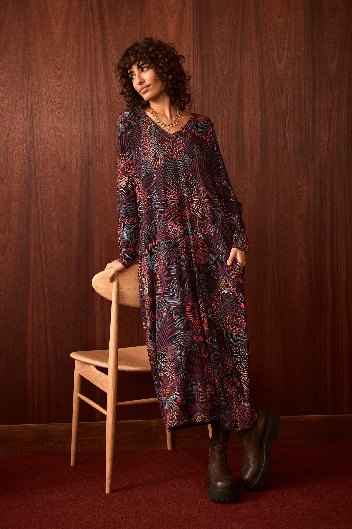 A person in the Abstract Linear Floral Jersey Bubble Dress and brown boots leans on a wooden chair, standing in front of a dark wood-paneled wall and red carpeted floor.