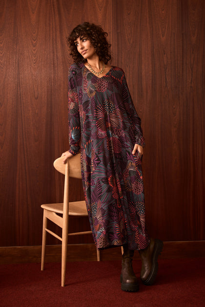 A person in the Abstract Linear Floral Jersey Bubble Dress and brown boots leans on a wooden chair, standing in front of a dark wood-paneled wall and red carpeted floor.