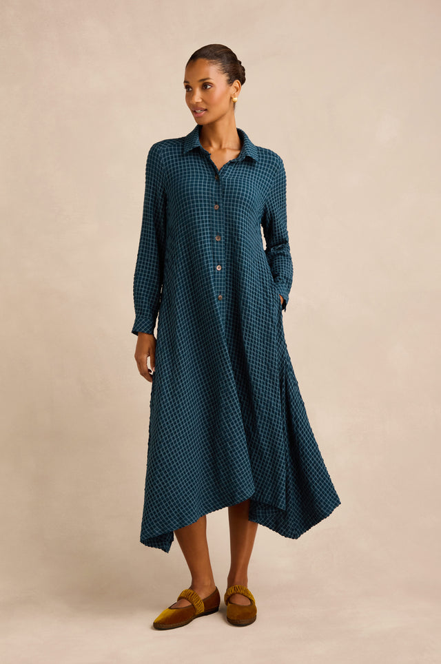 A woman stands against a plain beige background, looking to the side, wearing the Seersucker Squares Shirt Dress—a long-sleeve, teal checkered dress with an asymmetrical hem crafted from seersucker squares and made in England.
