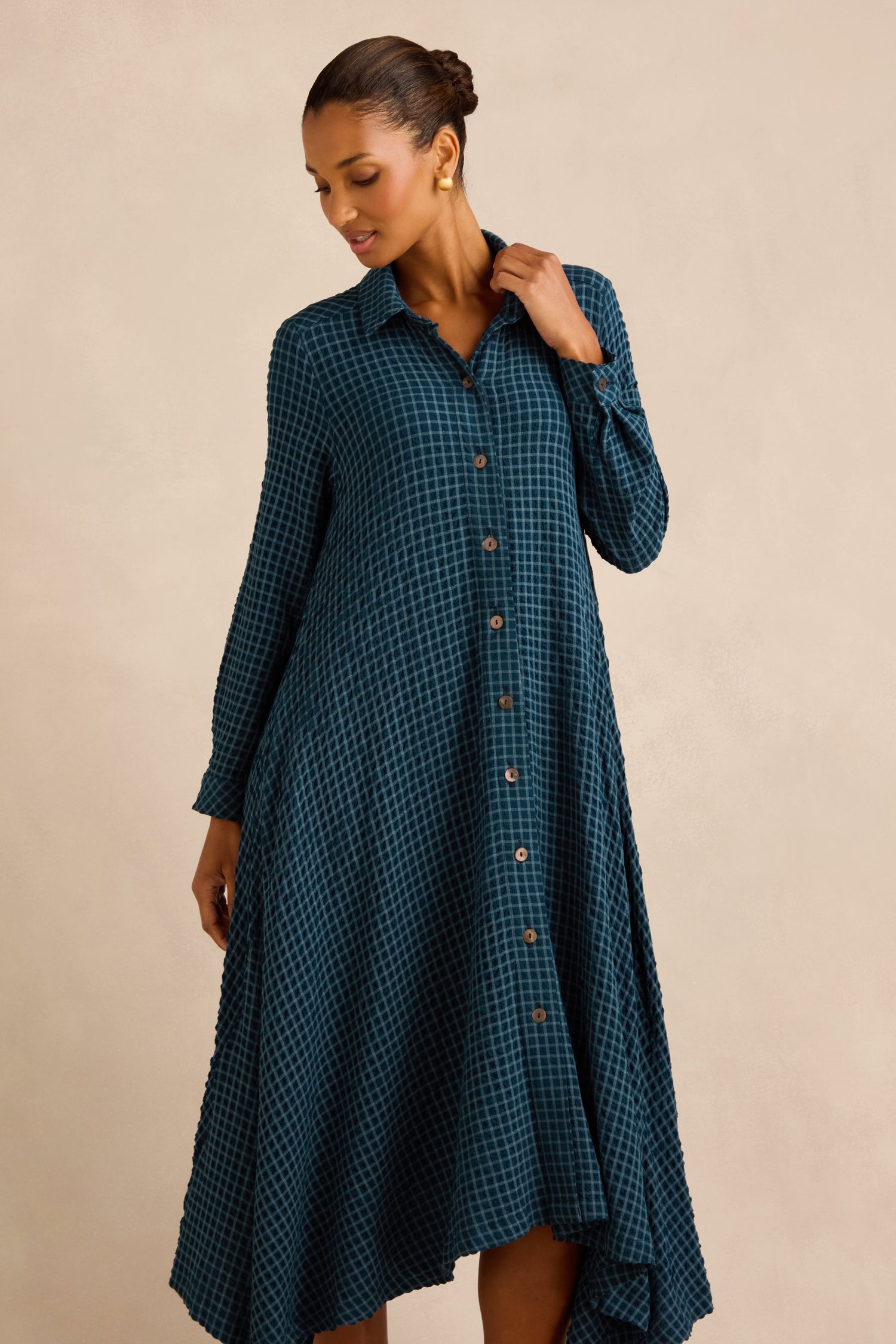 A woman wearing the Seersucker Squares Shirt Dress—a teal plaid, long-sleeved dress made in England—stands against a plain beige background, looking downward.