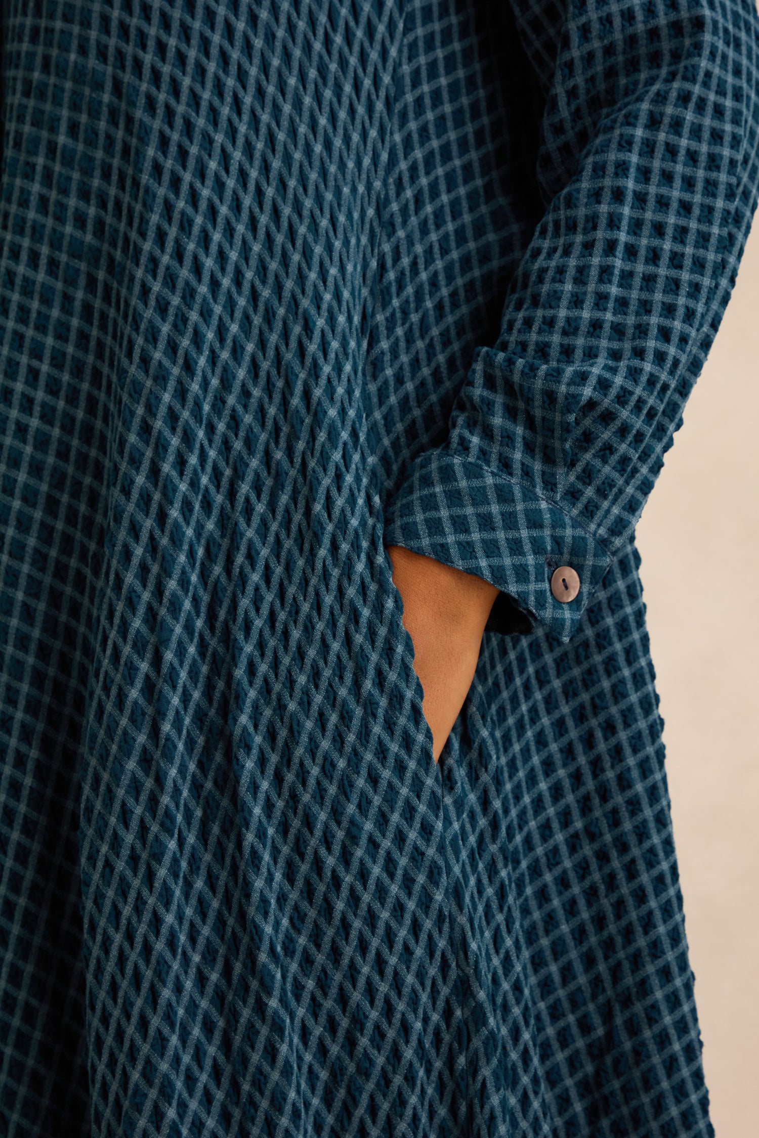 Close-up of a person wearing the Seersucker Squares Shirt Dress, featuring textured blue checkered fabric, with one hand in the side pocket and the sleeve cuff secured with a button.