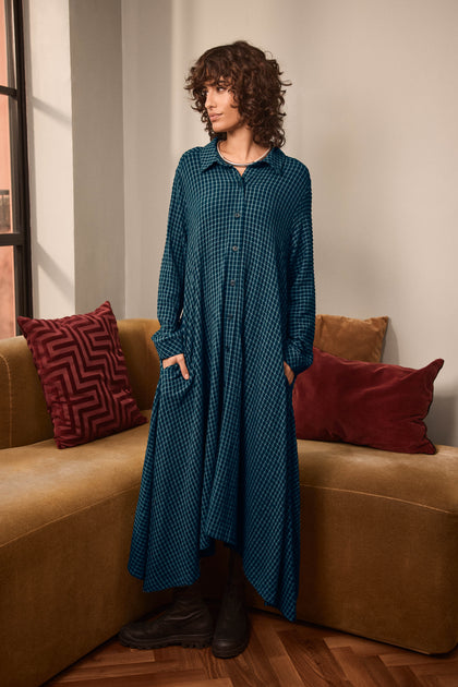 A person stands indoors on a tan sofa, wearing the Seersucker Squares Shirt Dress—a long, teal checkered design made in England—paired with black boots and hands tucked into pockets. Decorative pillows accent the sofa, and a window is visible in the background.