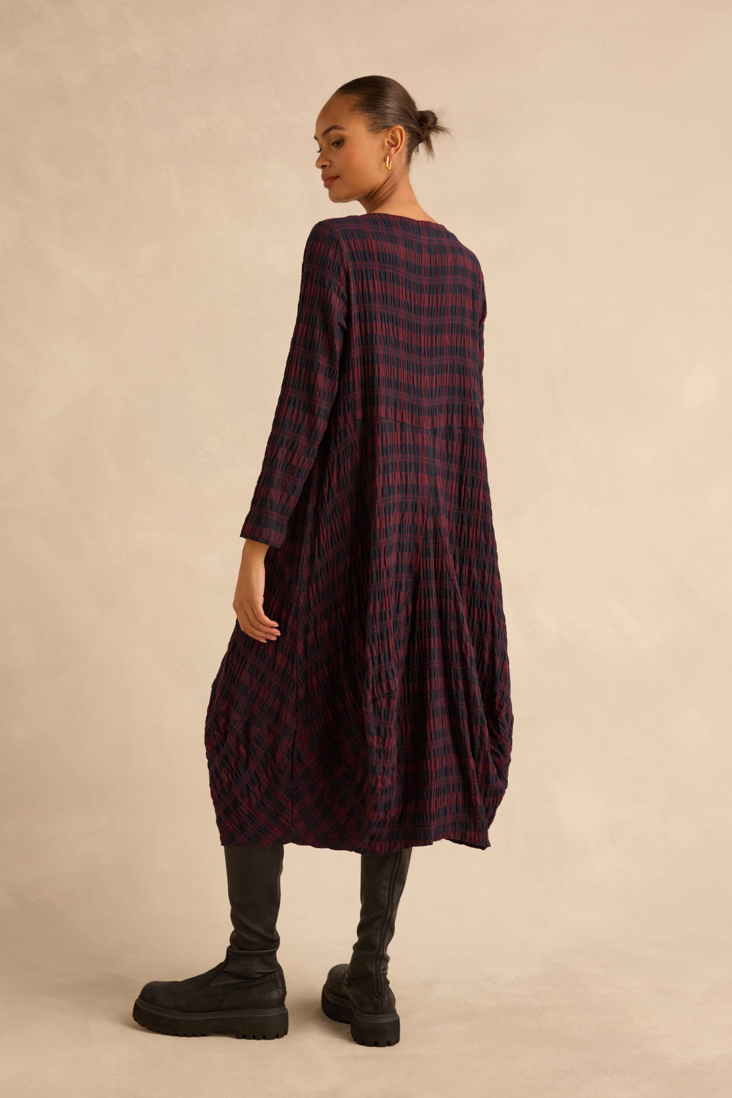 A person seen from the back wearing a Crinkle Check Bubble Dress, a long-sleeved, midi-length bubble skirt dress with a crinkled, dark red and navy check pattern, paired with black boots.