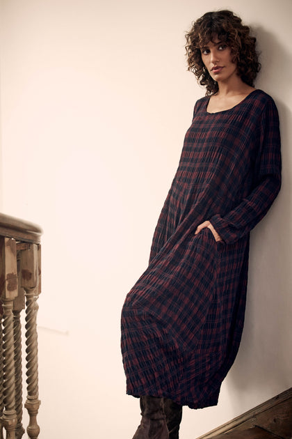 A woman leans against a wall wearing a long-sleeved, dark red and black Crinkle Check Bubble Dress.