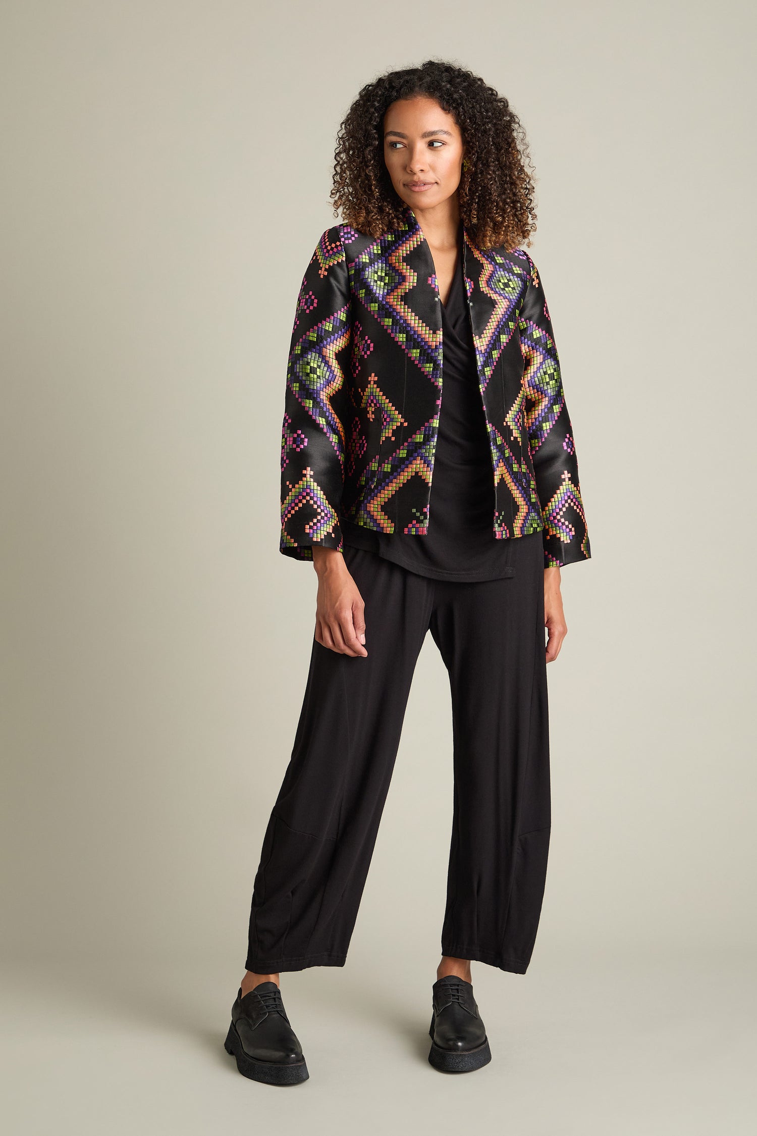 A person stands against a plain background, dressed in a sleek black outfit complemented by the Pixelated Jacquard Jacket featuring a vibrant, multicolored jacquard pattern, and black shoes.