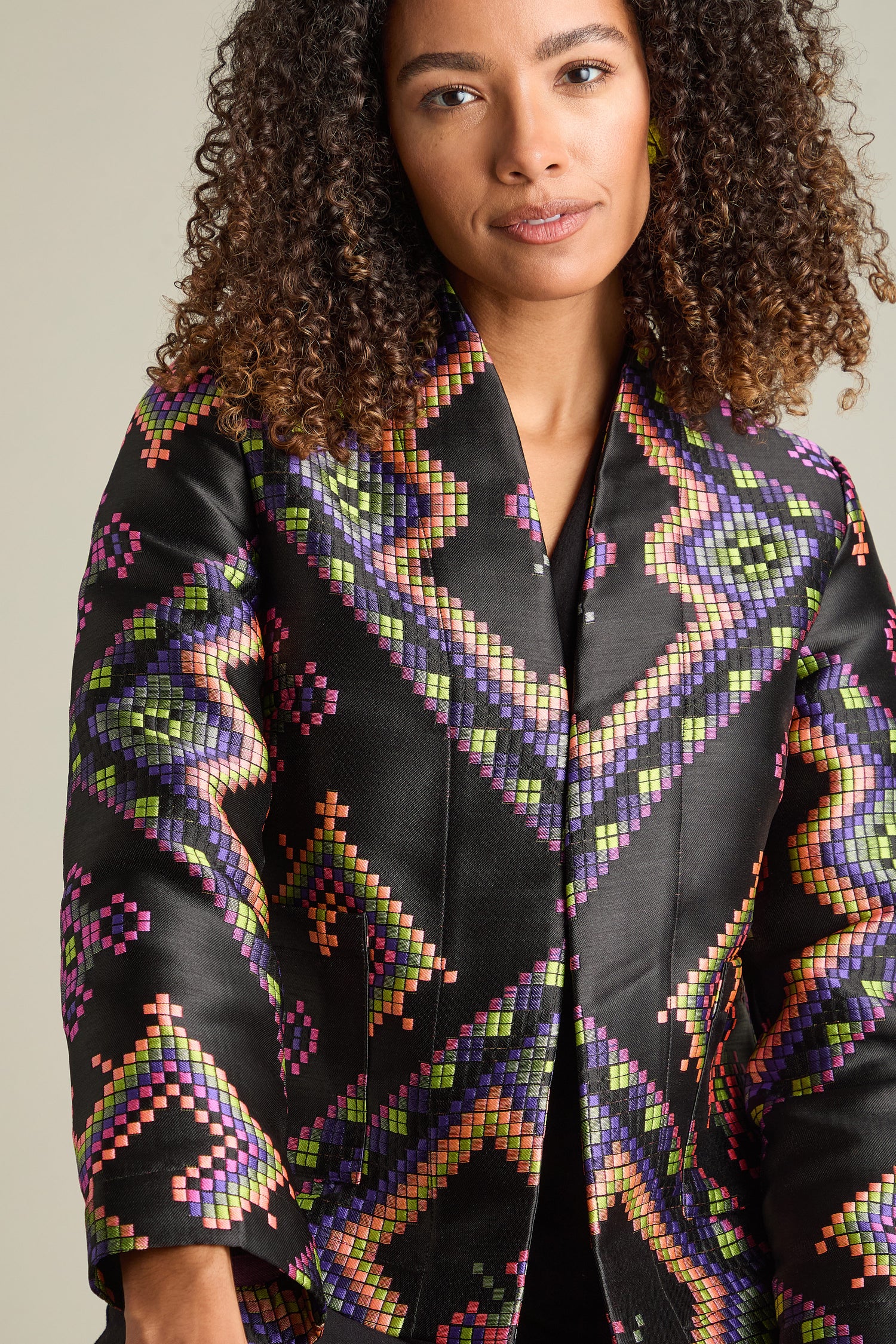 A person with curly hair models the Pixelated Jacquard Jacket adorned with vibrant geometric patterns, gazing slightly to the side in a strikingly modern pose.