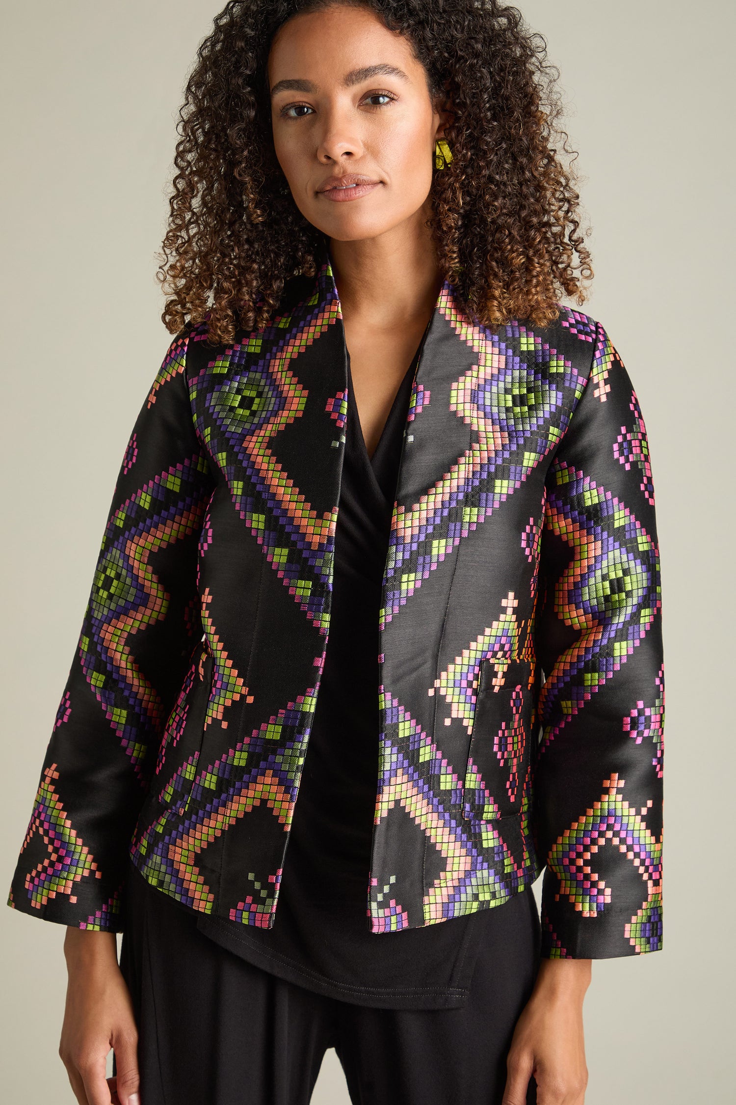 A person stands with curly hair, dressed in a black blouse, black pants, and a Pixelated Jacquard Jacket. The jacket boasts a contemporary look with its geometric design featuring green, purple, yellow, and pink in a multicoloured jacquard pattern.