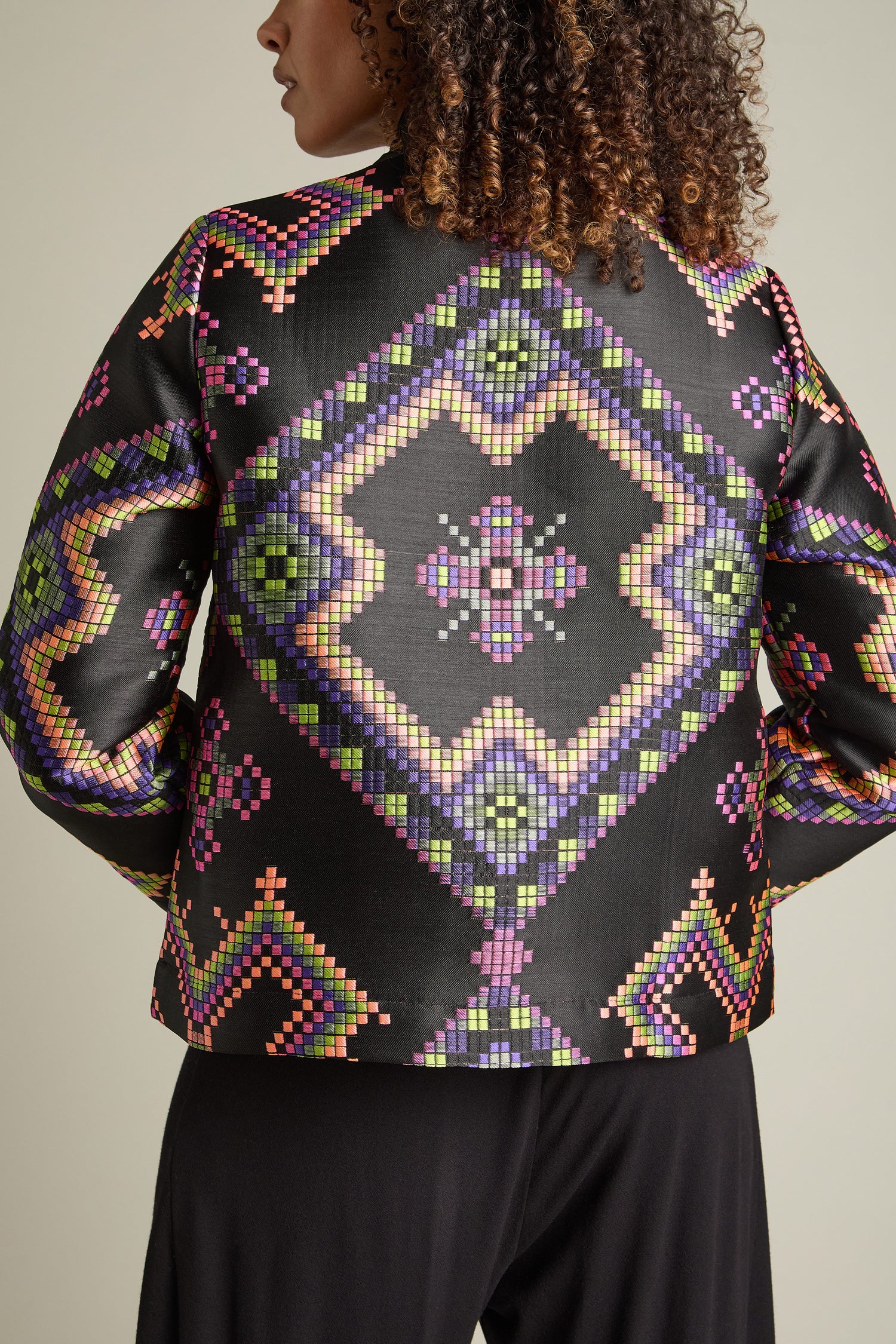 A person with curly hair is pictured from the back, donning a Pixelated Jacquard Jacket. The jacket is black with a vibrant and multicoloured jacquard pattern featuring modern designs that include diamond and cross shapes.