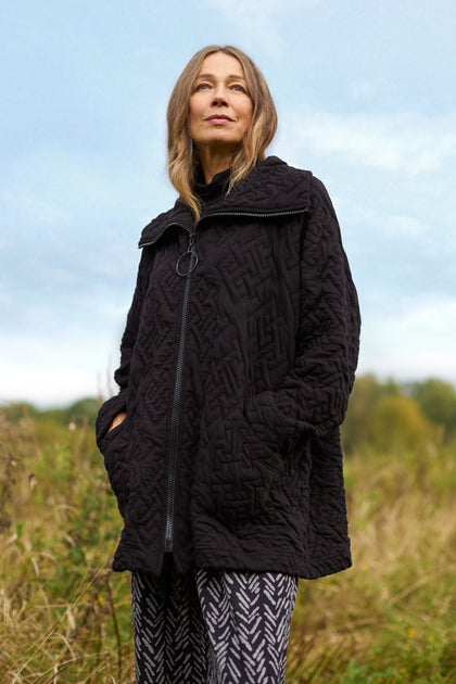 A woman stands outdoors wearing the chic Abstract Quilted Cotton Jacket, featuring dolman sleeves, paired with patterned pants. In the background, a grassy field extends under a cloudy sky, forming a peaceful scene.