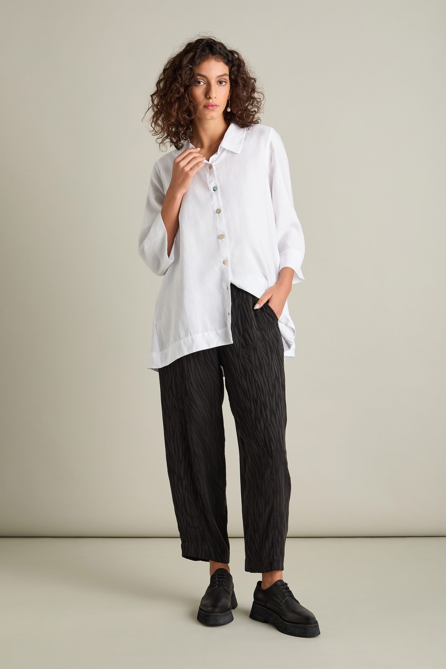 A person with curly hair is standing against a plain background, wearing a flowing white button-up shirt made of silky-feel fabric, the Abstract Lines Jacquard Bubble Trouser in black, and black shoes. The ensemble creates a striking contemporary look.