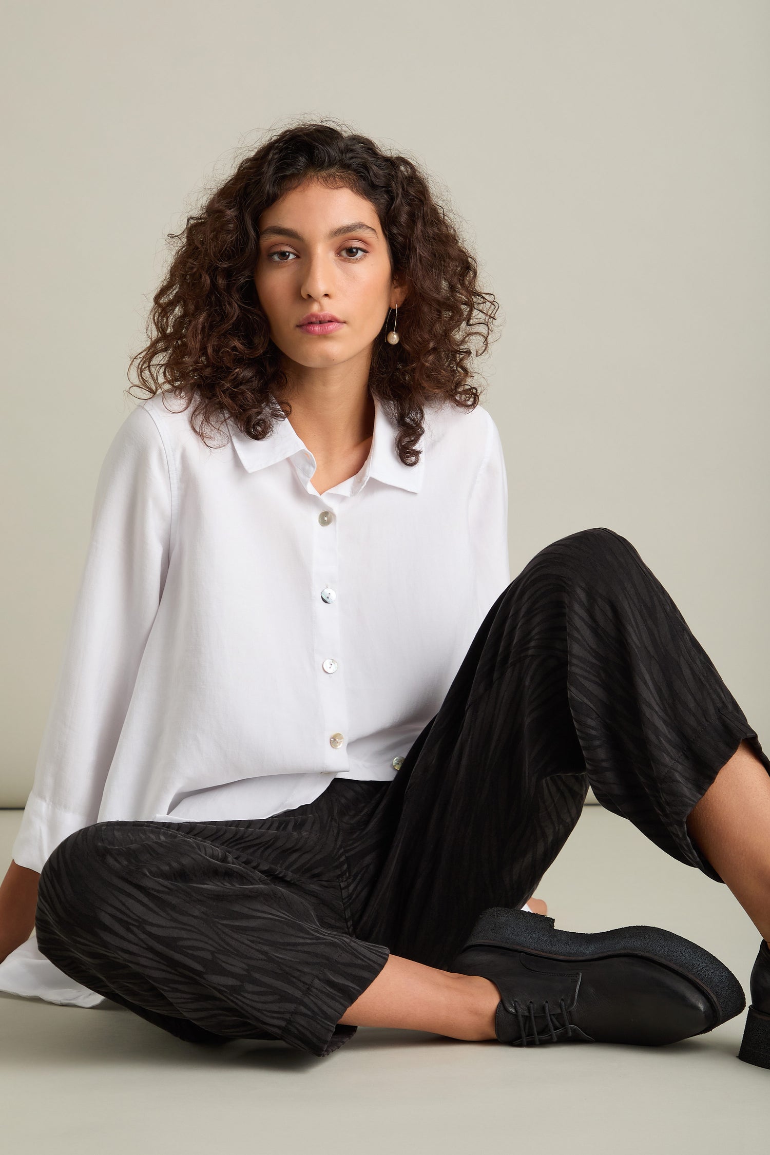 A person with curly hair is seated, wearing a white button-up shirt, Abstract Lines Jacquard Bubble Trouser, and black shoes, against a plain background for a contemporary look.