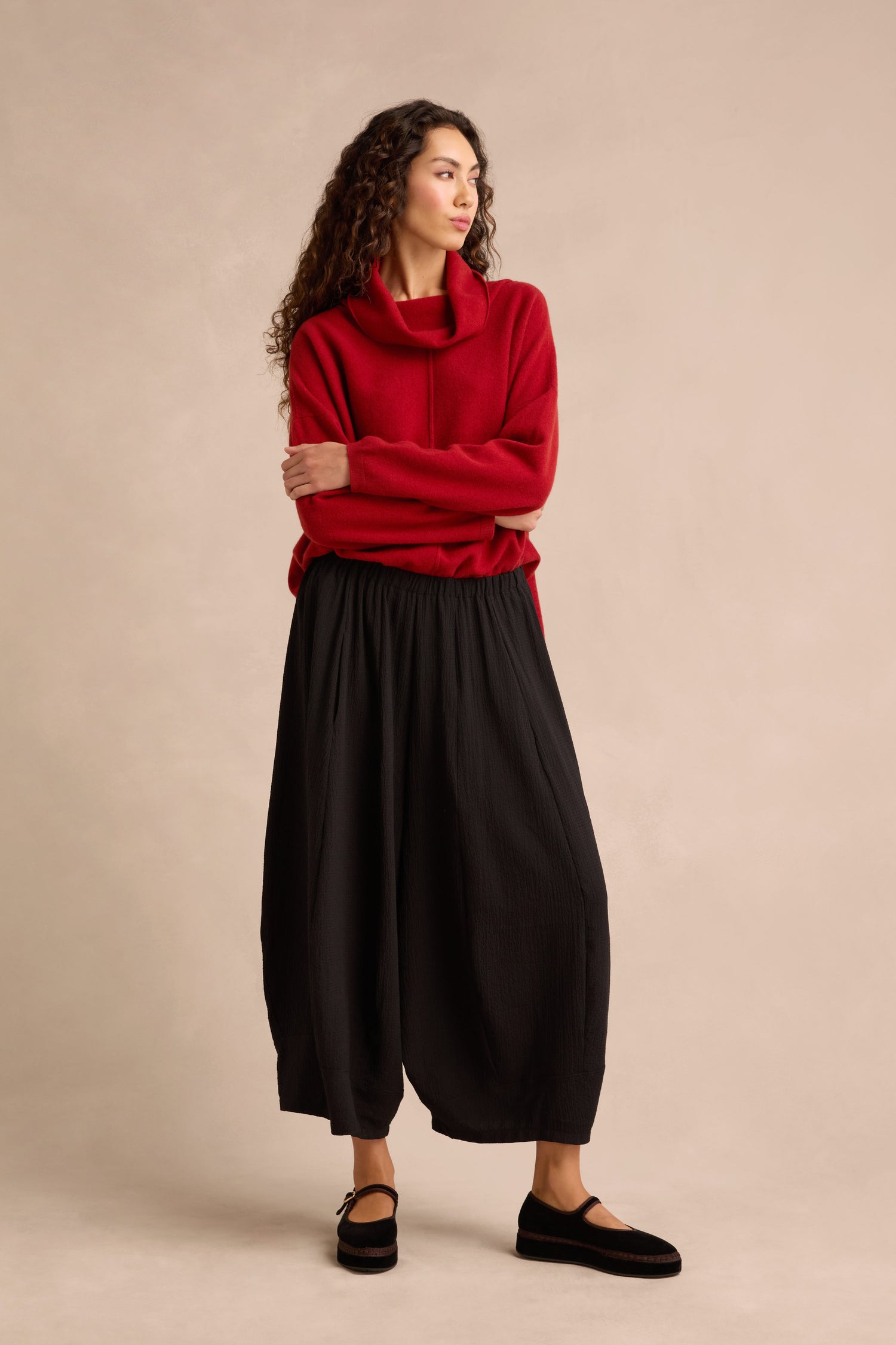 A woman stands with arms crossed, looking to the side against a plain beige background, wearing a red cowl-neck sweater and cropped Fine Crinkle Barrel Trousers in black, paired with black shoes.