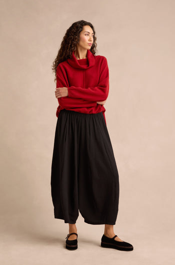 A woman stands with arms crossed, looking to the side against a plain beige background, wearing a red cowl-neck sweater and cropped Fine Crinkle Barrel Trousers in black, paired with black shoes.