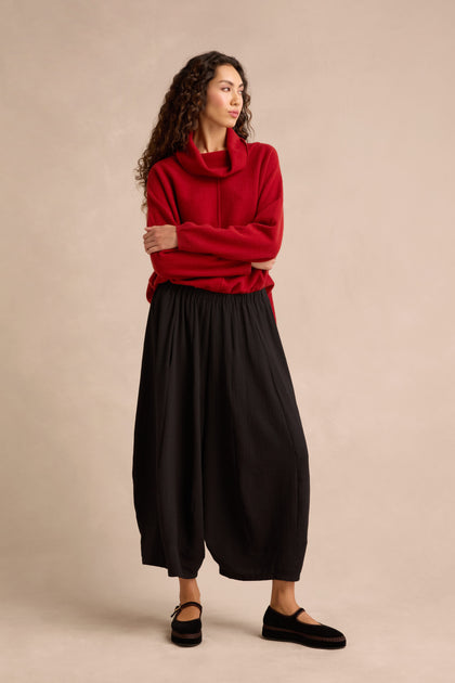 A woman stands with arms crossed, looking to the side against a plain beige background, wearing a red cowl-neck sweater and cropped Fine Crinkle Barrel Trousers in black, paired with black shoes.