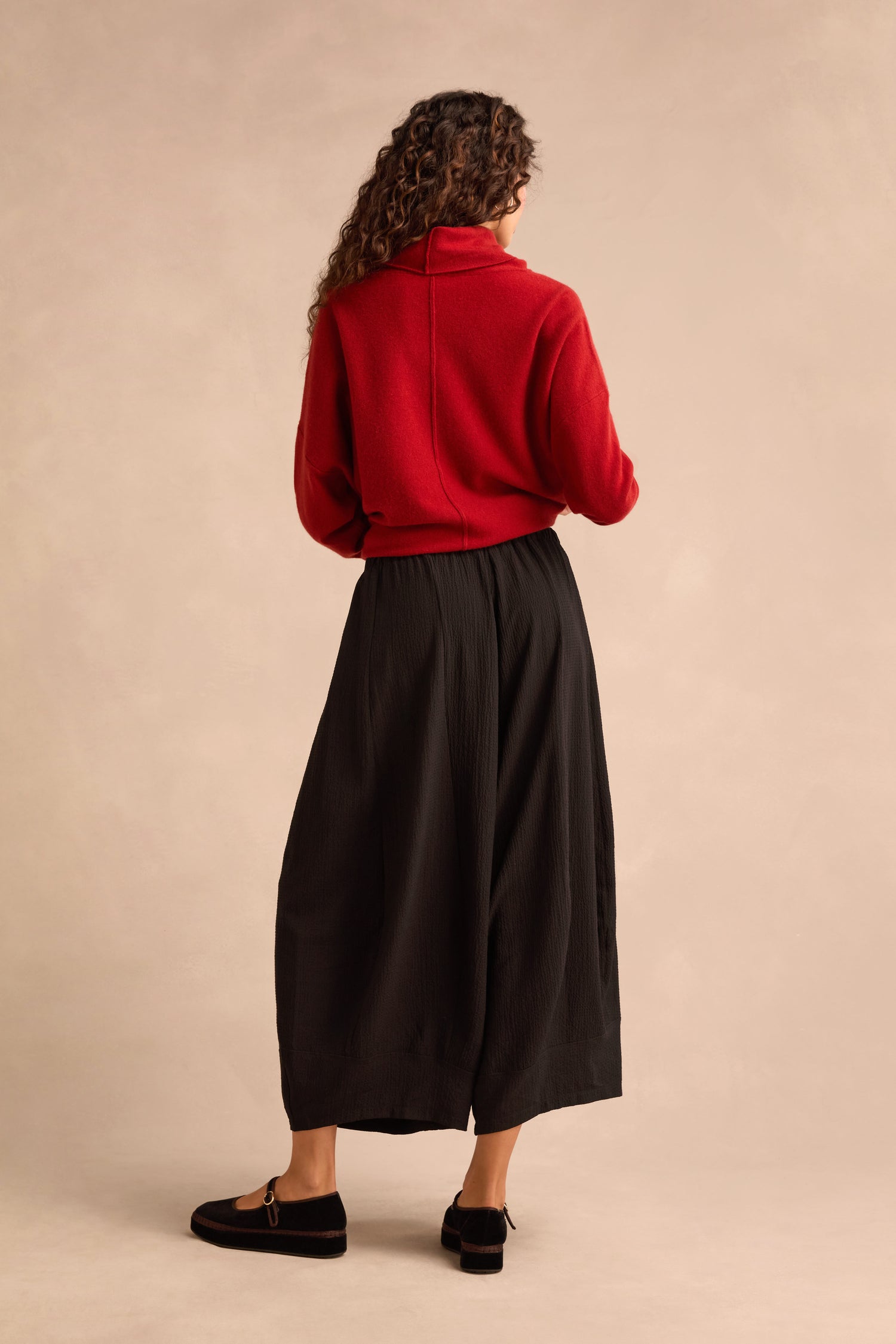 A person is standing with their back turned, wearing a red turtleneck sweater, Fine Crinkle Barrel Trousers in black with a cropped length, and black loafers, set against a neutral studio background.