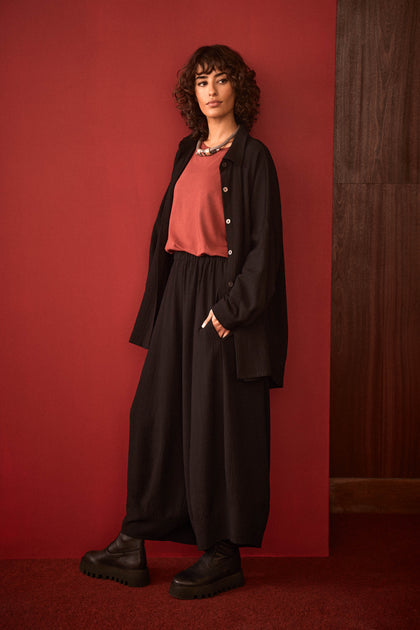 A woman with curly hair stands against a red wall, wearing a red top, black Fine Crinkle Barrel Trousers in a cropped length, a black jacket, and black platform shoes on a red carpet.