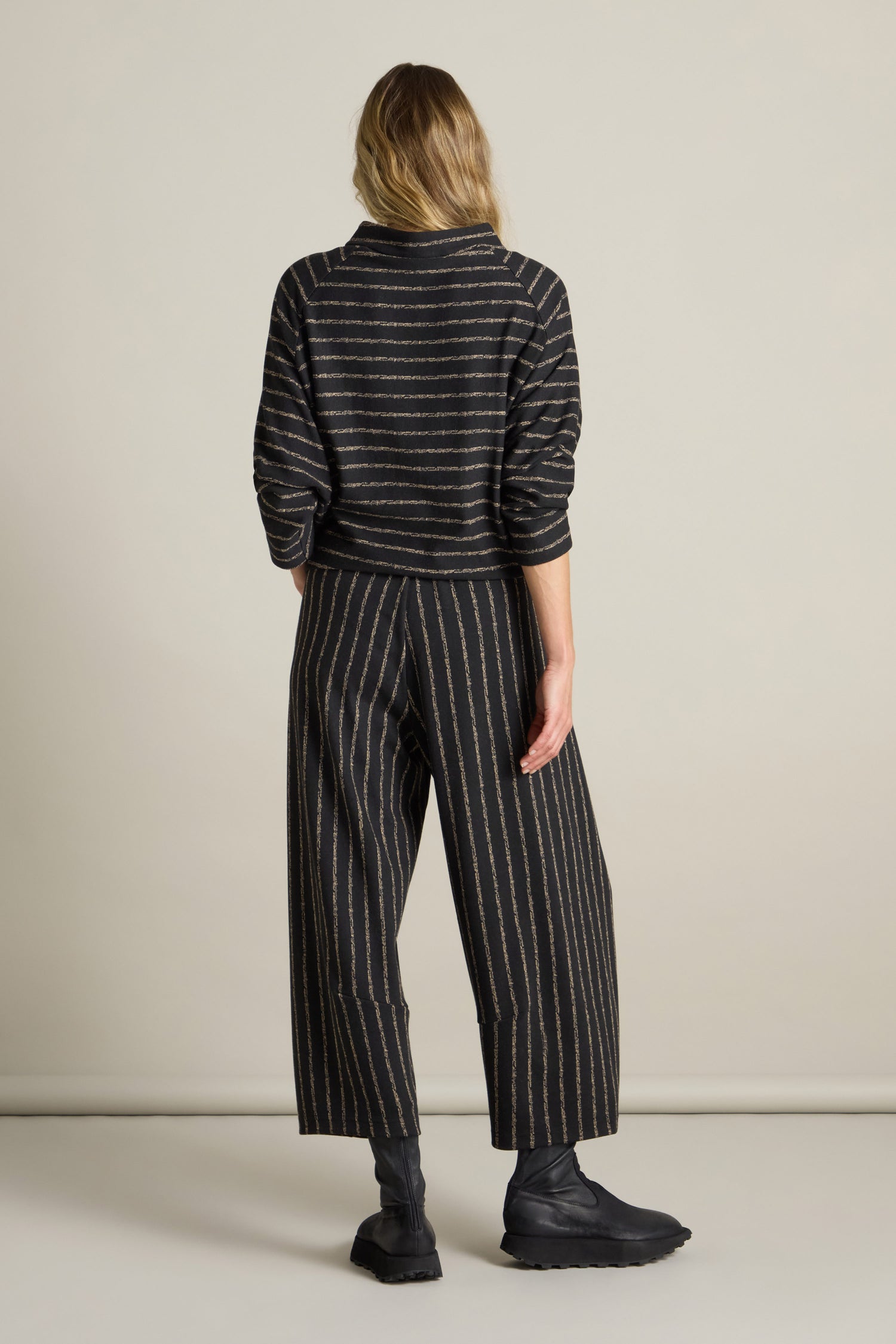 A person with long hair stands facing away, wearing a black, long-sleeved, striped shirt and matching Broken Stripe Jersey Bubble Trousers, complemented by black boots.