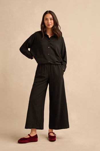 A woman stands against a beige background wearing a loose black button-up top, Mini Crinkle Viscose Wide Leg Trousers with an elasticated waist, and red slippers. One hand rests in her pocket; her hair is loose and wavy.