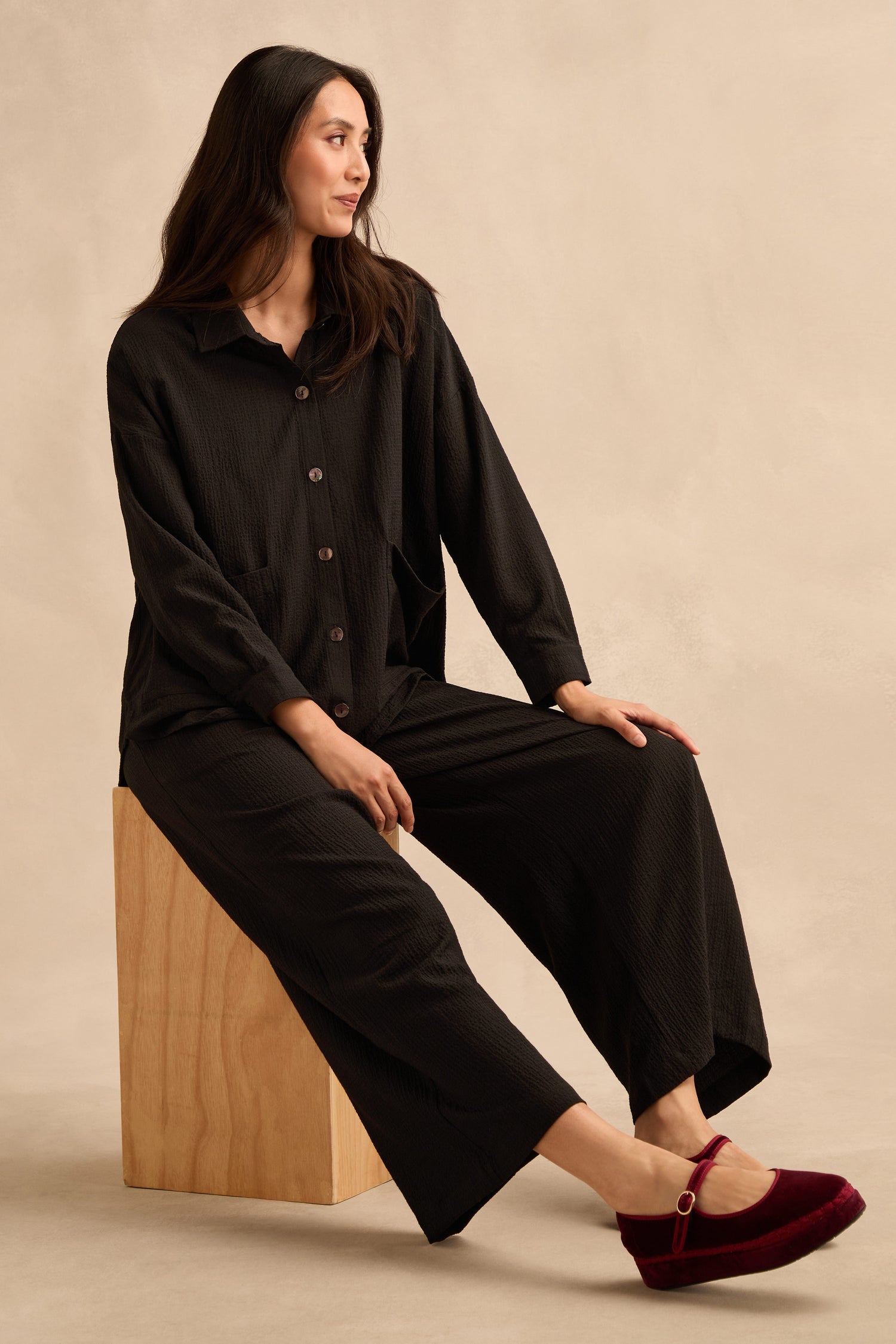 A woman sits sideways on a wooden block against a beige background, wearing a loose black button-up outfit paired with the Mini Crinkle Viscose Wide Leg Trouser, which features an elasticated waist, and completes the look with red velvet flats.