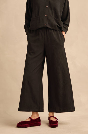A person wearing the Mini Crinkle Viscose Wide Leg Trouser in black with an elasticated waist, paired with a matching black button-up top and red velvet Mary Jane flats, stands against a beige background.