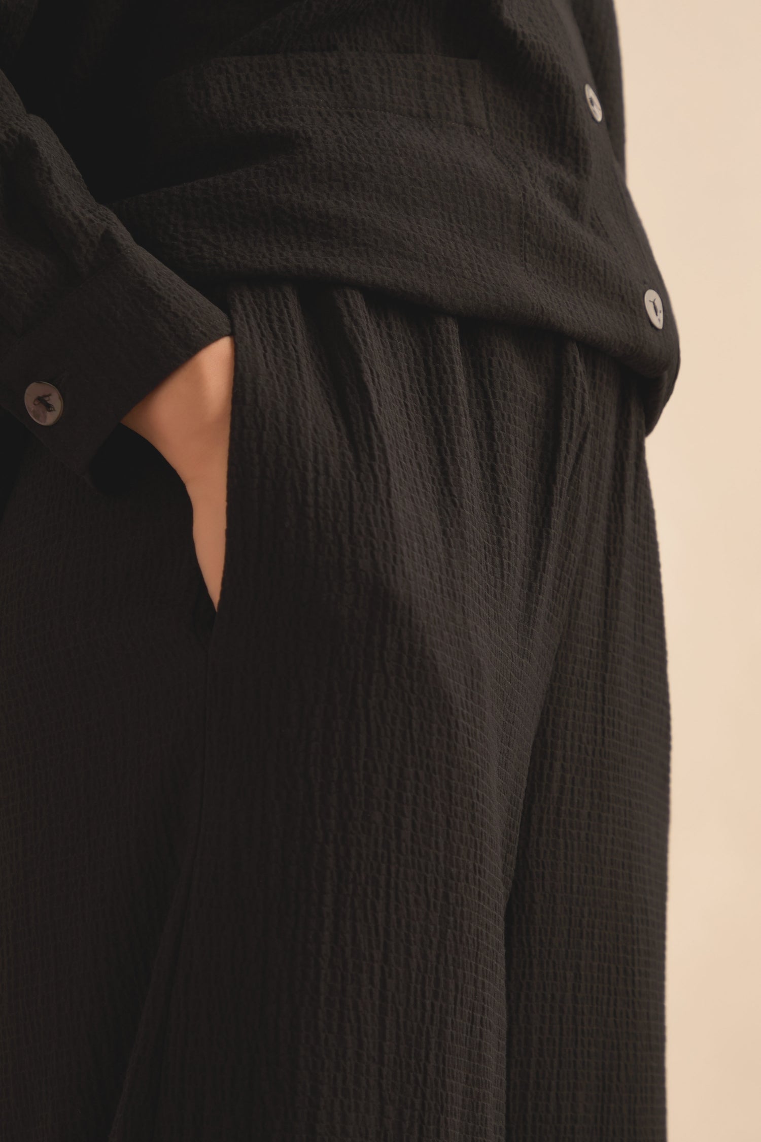A close-up shows a person wearing the Mini Crinkle Viscose Wide Leg Trouser in black, featuring an elasticated waist and their right hand tucked into the pocket. Only the torso and upper leg are visible.