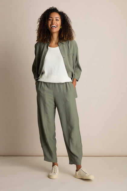 A person with curly hair smiles while wearing a light green button-up shirt and Textured Viscose Bubble Trouser with an elasticated waistband over a white top, hands in pockets, standing against a plain beige background.