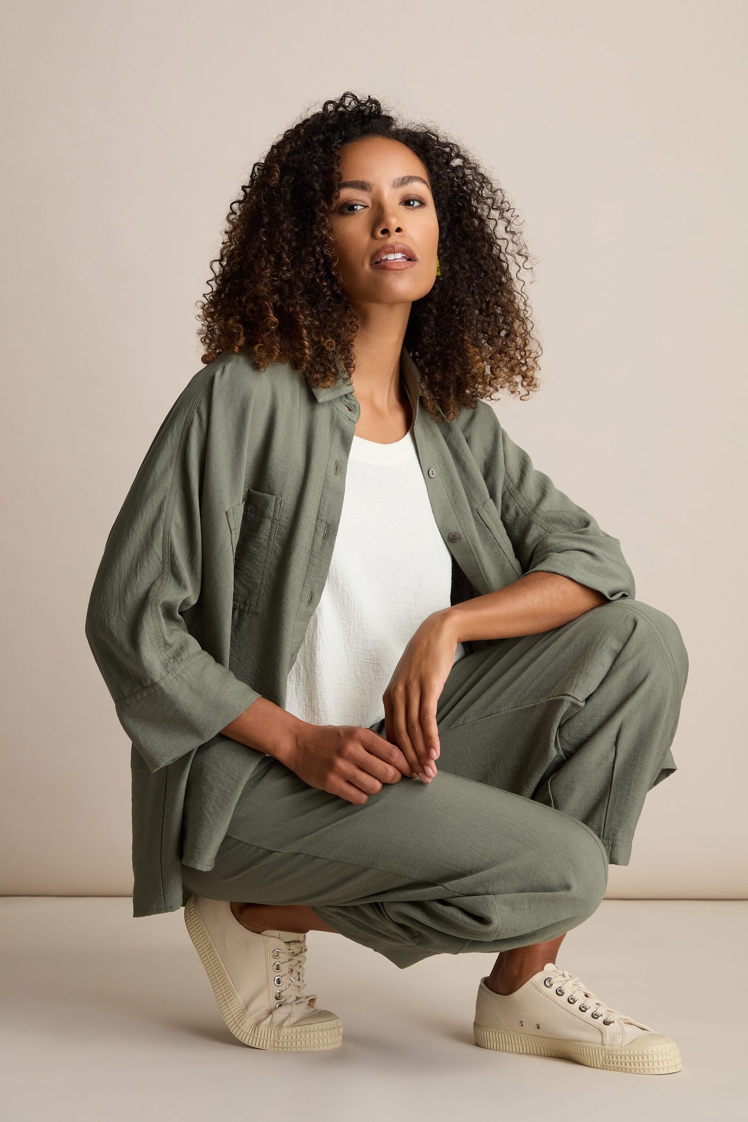 A person with curly hair crouches while wearing an olive green shirt, Textured Viscose Bubble Trousers with an elasticated waistband, a white top, and beige shoes.