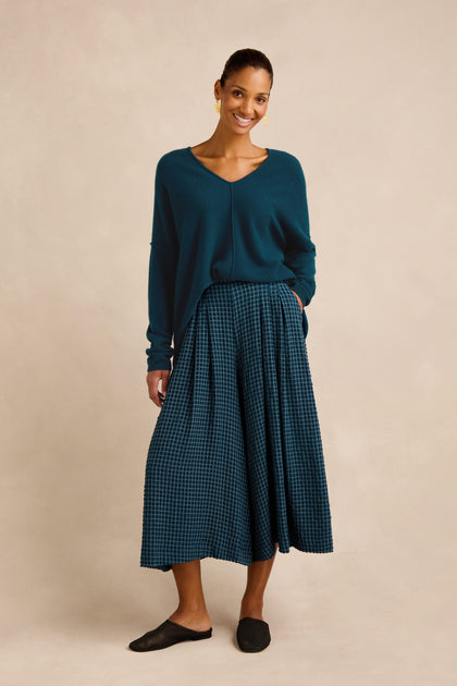 Woman standing and smiling, wearing a teal V-neck sweater, Seersucker Squares Wide Trousers in a teal checkered pattern, black flat shoes, and gold earrings against a neutral background.