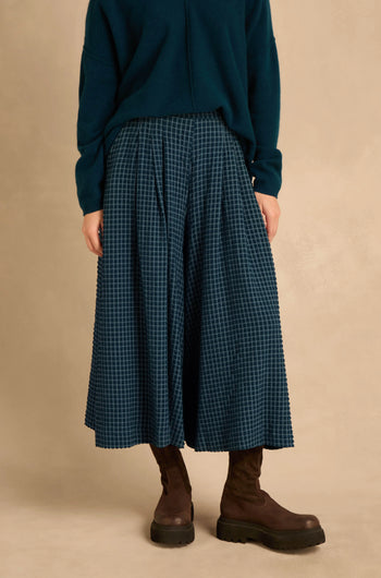 A person wears a dark teal long-sleeved sweater, Seersucker Squares Wide Trousers with a blue and black grid pattern, and dark brown ankle boots.