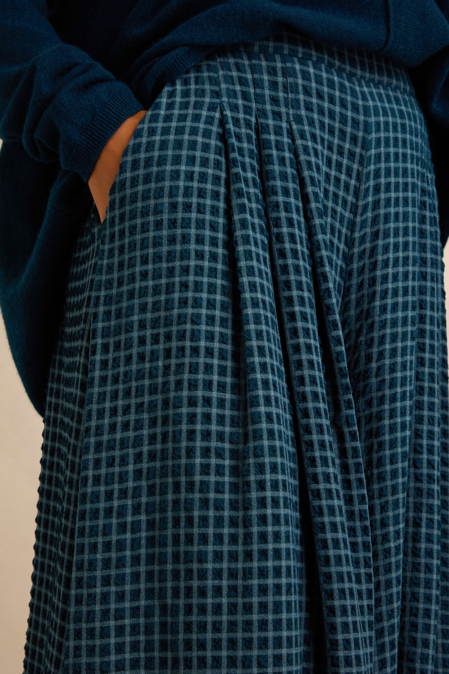 Wearing a dark blue sweater and a textured blue plaid skirt with one hand in the pocket, this sophisticated look can be easily elevated by pairing it with our Seersucker Squares Wide Trousers for added style versatility.