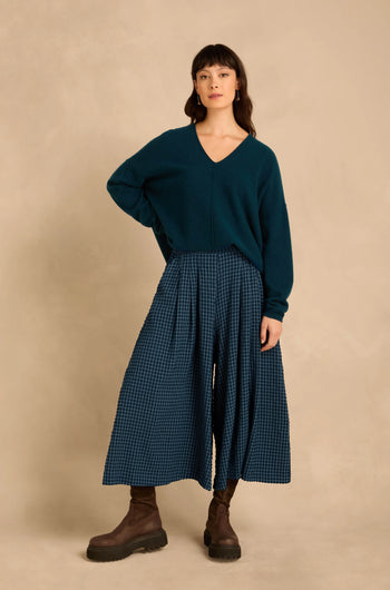 A model wears a deep teal V-neck sweater, Seersucker Squares Wide Trousers in a blue checkered pattern, and brown ankle boots.