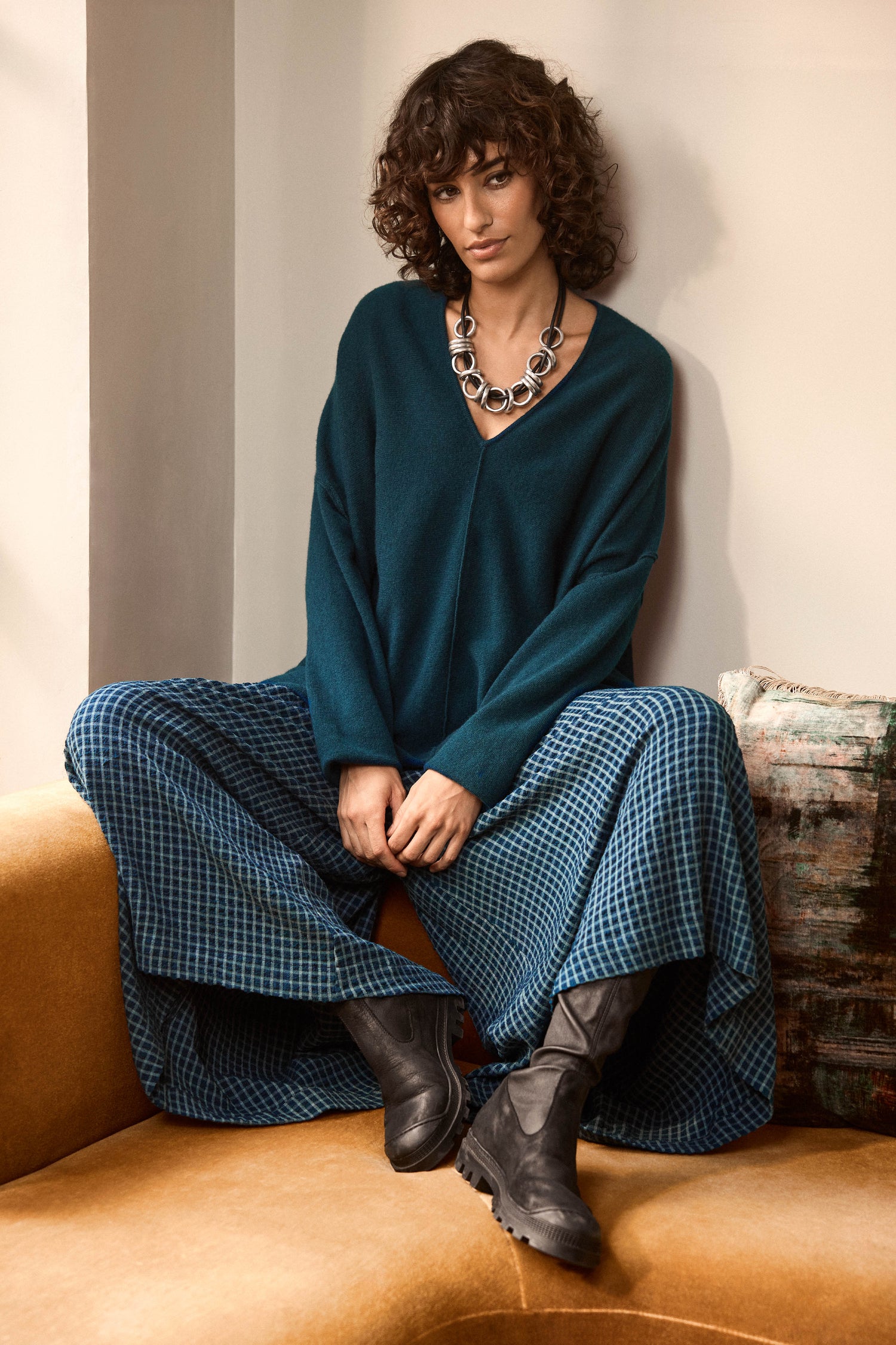 A person with curly hair sits on a tan couch, wearing a teal sweater, Seersucker Squares Wide Trousers, black boots, and a chunky silver necklace.