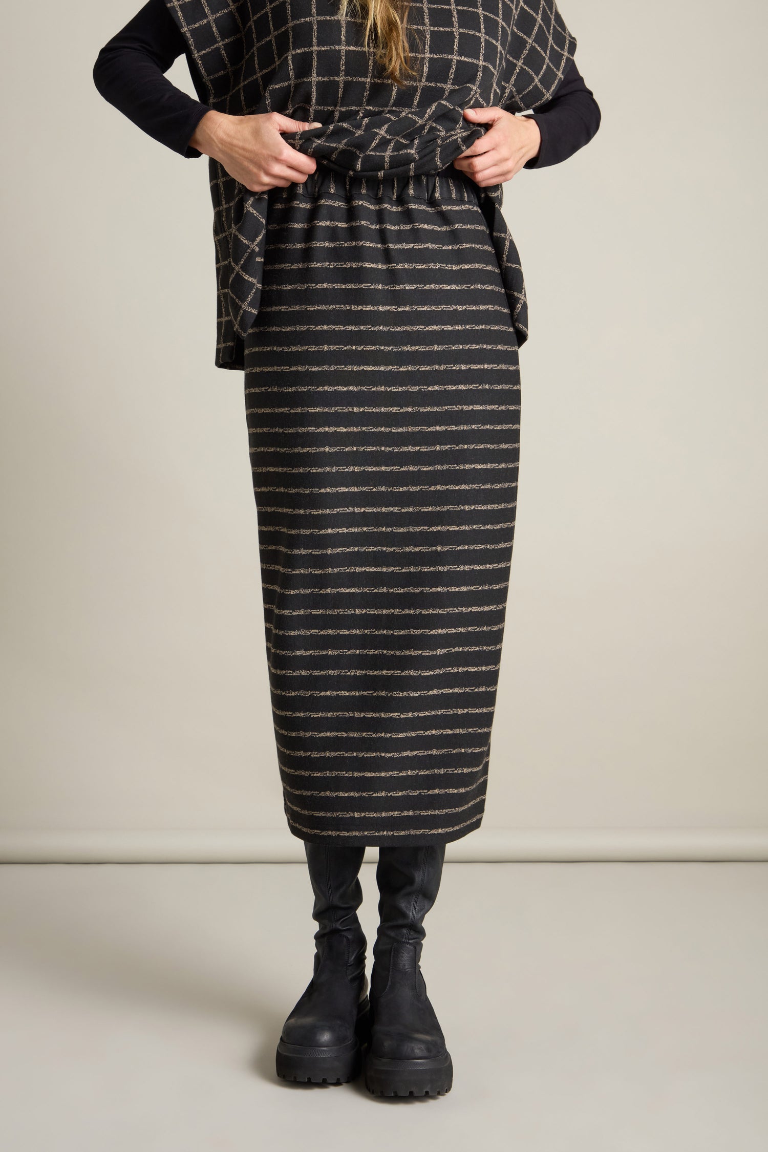 A person clad in a knee-length, dark-colored Broken Stripe Jersey Tube Skirt, paired with black leggings and boots. Crafted from soft jersey fabric, this slim-fit tube skirt enhances their style as they adjust it against a light background.