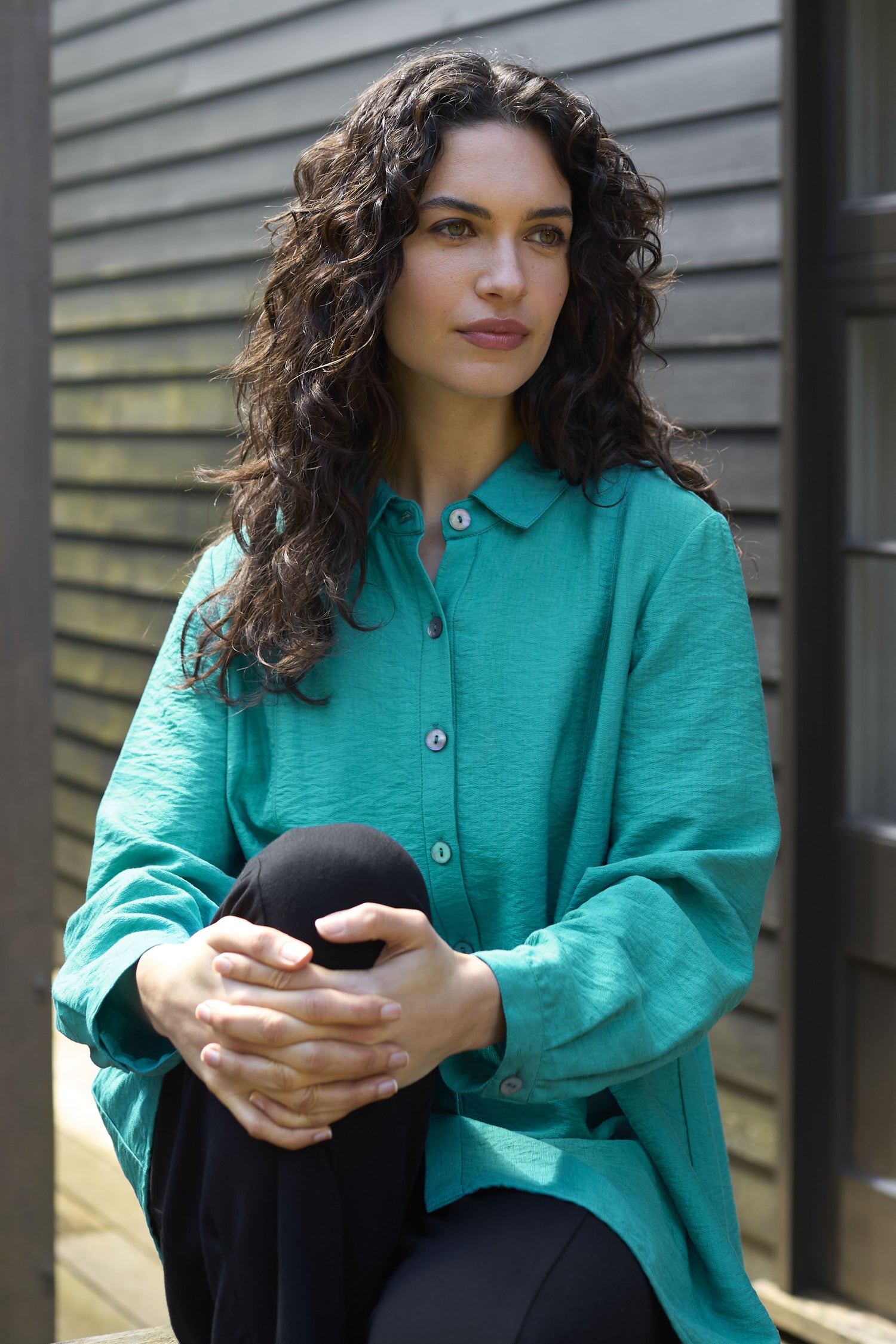 A person with long curly hair wearing a green Textured Viscose Pleat Shirt and turquoise blue Bubble Trousers sits outdoors with hands clasped over their knees.