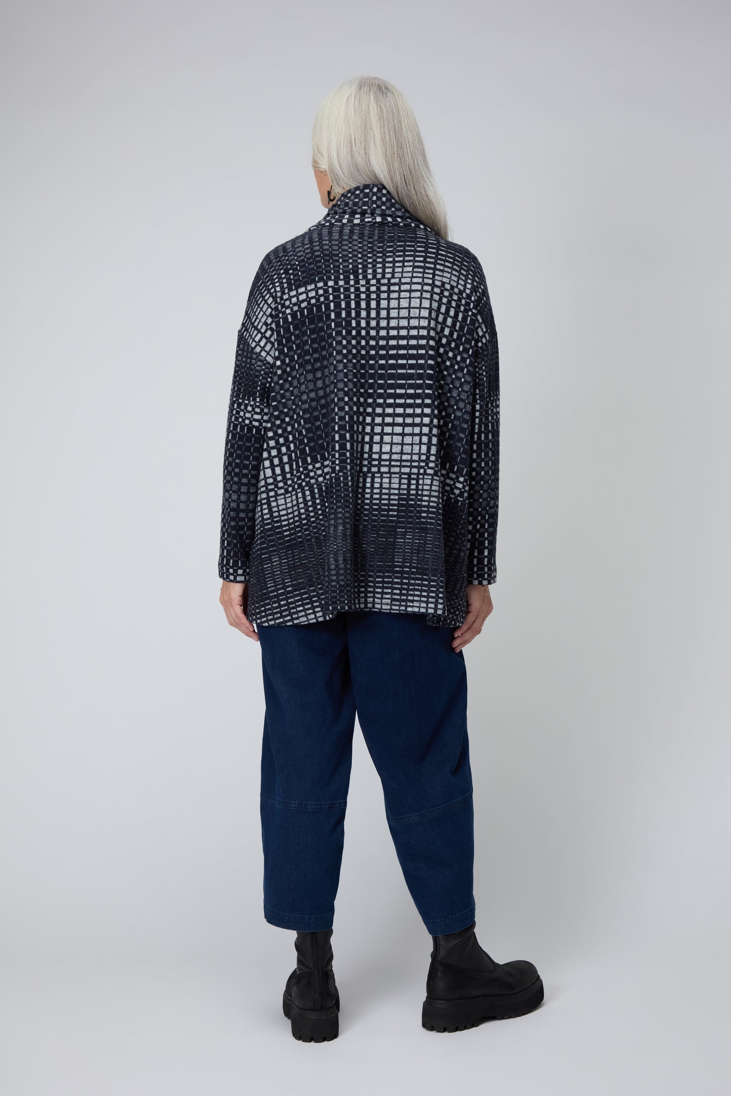 A person with long gray hair stands with their back to the camera, wearing the Illusion Check Jersey Tunic, dark blue pants, and black boots.